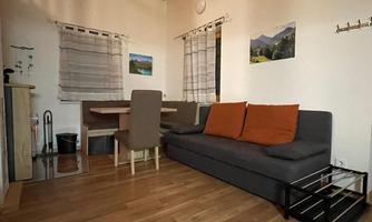 A cozy room with a gray sofa and orange cushions. In the background, there is a dining table and decorative pictures on the wall.