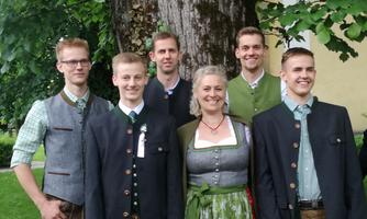 A group of six people in traditional attire stands in front of a large tree. The men are wearing short leather pants and the woman a festive dirndl.
