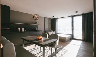 A modern living room with a dining area, elegant furniture, and large windows. It offers plenty of light and a view outside.