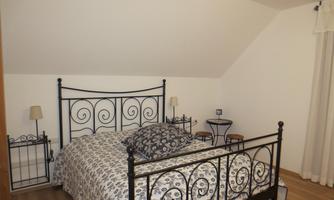 A cozy bedroom with a wrought-iron bed and decorative cushions. The walls are light and the floor is wooden, creating a warm atmosphere.