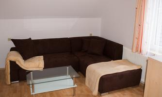 A cozy brown couch with a glass side table. The room has light walls and large windows with curtains.