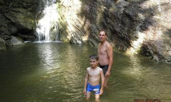 A father and his son are standing in the water of a small pool in front of a waterfall. The surroundings are characterized by rocks and natural vegetation.