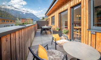 A cozy terrace with wooden walls and chairs. In the background, there are mountains and a clear sky.