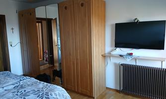 A bedroom with a large bed, a wood-colored wardrobe, and a television on the wall. The floor is made of wood, and there is a radiator in the corner.