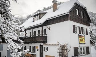 A charming house in the snow with a traditional design and a balcony. The surroundings are wintry with snow-covered trees.