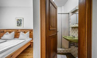 A simple room with a wooden bed and two pillows. In the background, a bathroom with a shower and sink can be seen.