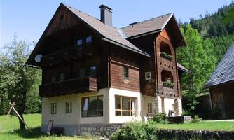 A traditional wooden house with several balconies in a green environment. Trees and hills can be seen in the background.