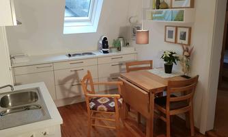 A bright, modern kitchen with a small dining table set. The window provides plenty of natural light, and there are decorative plants.