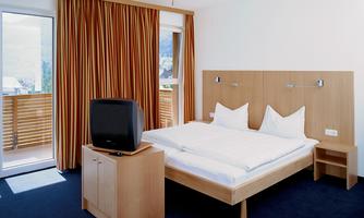 A simple hotel room with a double bed and two pillows. There is a television and large windows with curtains.