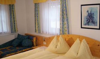 A cozy bedroom with a large double bed and colorful cushions. The windows are adorned with bright curtains and a picture hangs on the wall.