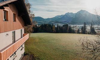 A charming house with wood cladding, surrounded by a green meadow. In the background, impressive mountains and a clear sky can be seen.
