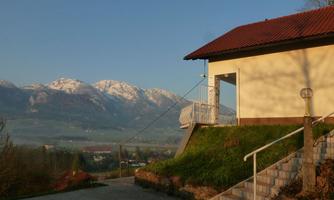 A modern house with a red roof and a staircase stands on a hill. In the background, snow-covered mountains and a clear sky are visible.