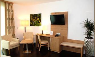 A modern hotel room with two comfortable chairs, a table, and a television. The lighting is warm, and a green piece of art hangs on the wall.