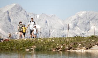 A family is hiking along a path with mountains in the background. In the foreground, a calm body of water and green meadows can be seen.