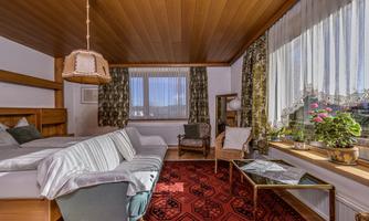 A cozy room with a wooden floor and a large window. In one corner stands a small table with plants, and there are comfortable furnishings.