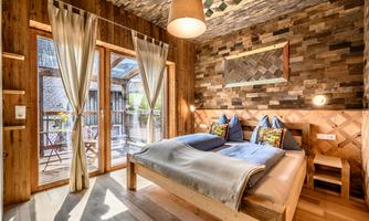 A cozy bedroom with wooden interiors and large windows. The bed is decorated with soft blankets and pillows, and there is a small balcony with a view outside.