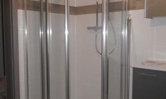 A modern shower area with transparent glass swing doors and wall cladding in neutral colors. The shower is equipped with a hand-held showerhead and is located in a bright bathroom.