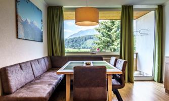 A modern dining room with a cozy bench and a table. Large windows offer a view of the mountains and nature.