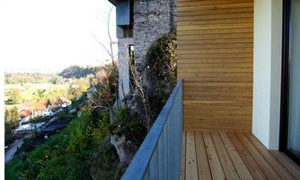 A balcony with a wooden floor and modern railings, surrounded by nature. In the background, landscape and buildings are visible.