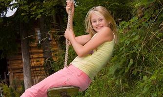 A girl is happily swinging on a rope in nature. In the background, trees and a wooden cabin can be seen.