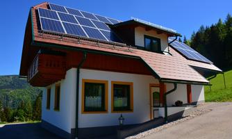 A modern house with solar panels on the roof. The surroundings are green and surrounded by trees.