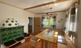 A bright, inviting dining room with a large wooden bench and a table made of wood. The windows let in a lot of light and a green tiled stove enhances the cozy atmosphere.