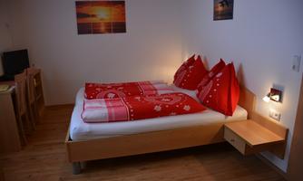 A cozy bedroom with a double bed and red pillows. Pictures hang on the wall, and there is a small table with a television.