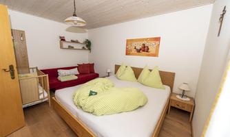 A cozy room with a double bed and a red couch. There is a picture on the wall, and there is a small cradle for a child.