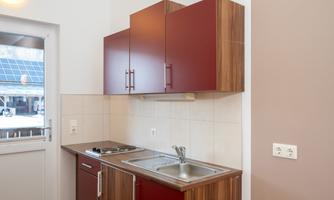 A modern kitchen with red upper cabinets and wooden shelves. The sink is placed next to a small cooking hob.