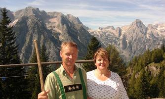 A man and a woman stand in front of an impressive mountain landscape. The surroundings are green with tall mountains in the background.