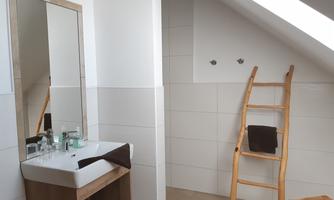 A modern bathroom with a sink, a mirror, and a wooden ladder. The walls are covered with white tiles, and there is a shower in the corner.