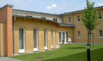 A modern building with a yellow facade and large windows. The outdoor area is green with a small tree and lawns.