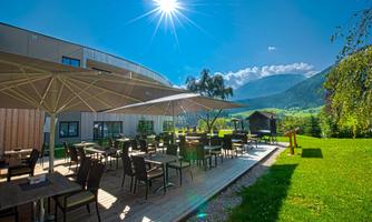 A sunny terrace with many tables and chairs. In the background, green meadows and mountains are visible.