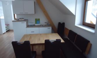 A modern, bright dining room with a rectangular wooden table and dark chairs. In the background, there is a functional kitchen unit with white cabinets.