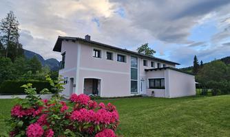 A modern, pink house surrounded by green grass and colorful flowers. The sky is partly cloudy and the landscape is tranquil and inviting.