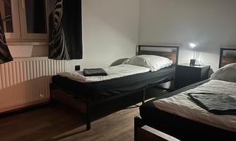A simple room with two single beds and a nightstand. The light is dim and there is a window with curtains.
