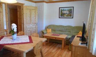 A cozy living room with a green sofa, a wooden table, and chairs. The walls are decorated with wood paneling and a picture.