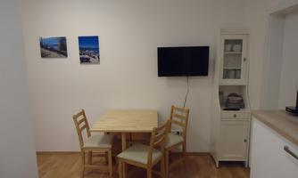A cozy dining area with a wooden table and four chairs. Two pictures hang on the wall and a television is installed.