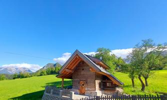 A cozy wooden house stands amidst a green meadow. The blue sky and the mountains in the background create a beautiful landscape.