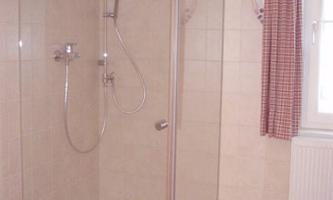A modern shower with a glass wall and tiled floor. The room has light walls and a window with a curtain.