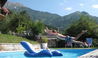 A boy jumps into the swimming pool and flies through the air. In the background, green mountains and beautiful trees can be seen.