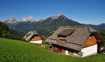 Two traditional wooden houses stand on a green meadow. In the background, majestic mountains and a clear blue sky can be seen.