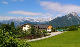A picturesque landscape with a farmhouse and tall mountains in the background. The sky is blue and the meadows are green.
