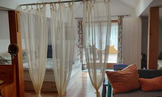 A cozy room with wooden flooring and a privacy screen made of white curtains. The windows bring a lot of light into the room, which is decorated with a sofa and cushions.
