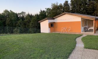 A modern house with an orange facade is situated in a green landscape. Next to the house, there is a small seating area.