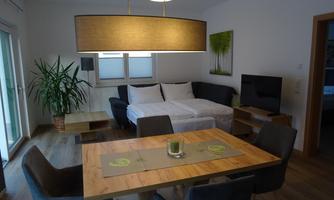 A modern living room with a sofa bed and a dining table. A large lampshade and a plant create a pleasant atmosphere.