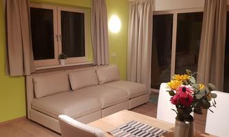 A modern living room with a beige sofa and a table with flowers. The walls are painted in a light green color and there are large windows with curtains.