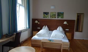 A cozy bedroom with a double bed and two pillows. The room has light walls, a small table, and large windows with curtains.