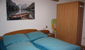 A cozy bedroom with a double bed and blue bed covers. Behind the bed hangs a picture of a boat on a calm water.