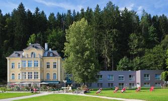 An elegant, historic building stands next to modern accommodations in a quiet setting. Surrounded by trees and a well-maintained green space, it invites relaxation.